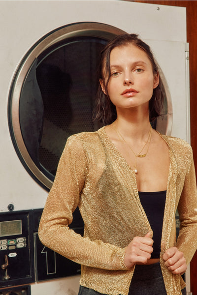a model wearing an open front cropped cardigan with long sleeves in a semi-translucent gold lurex knit. It has ribbed cuffs and hems. Shown from the front