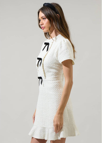 model wearing white textured tweed woven mini dress with mock neck, short sleeves with slightly puffed shoulders, bodice with vertical ruffled bib style and three black bows running down the middle. Dress has a-line skirt ending above the knee in a wide gathered ruffle. Shown from the side