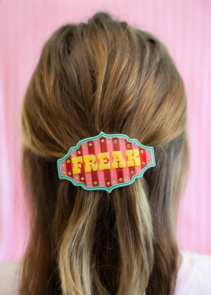 large cellulose acetate barrette in the shape of a circus tent banner with red and pink vertical stripes and blue and green border and "FREAK" in stylized yellow letters surrounded by a row of yellow rhinestones. Shown in updo of model from back
