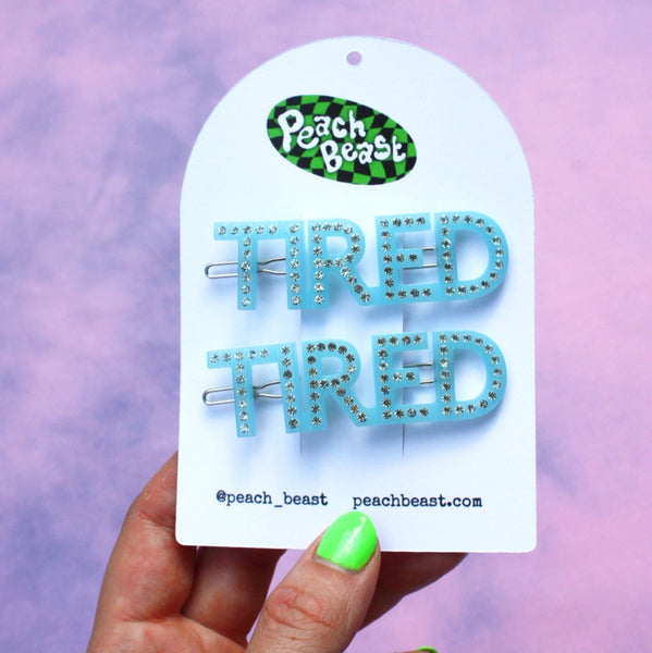pair of acetate baby blue barrettes spelling the word TIRED with silver rhinestone embellishments. Shown on packaging card