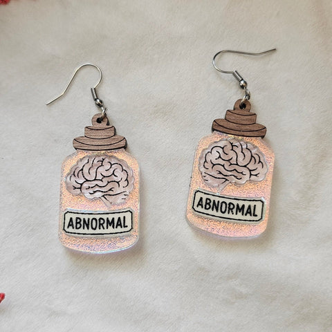 Dangle earrings shaped like jars with acrylic glitter “glass” and a wood lid with brains inside and 'Abnormal' labels