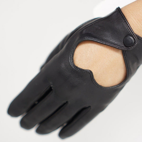 A pair of black faux leather wrist-length gloves with a heart-shaped cutout on the back of the hand and single snap closure. Shown worn by model in close up