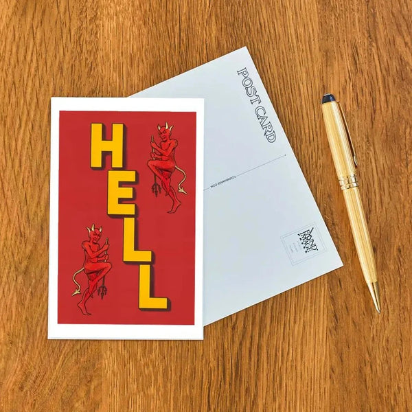Postcard with 'HELL' text and devil illustrations on a wooden surface with a pen.