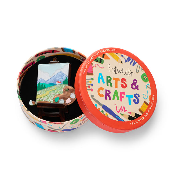 layered acrylic resin brooch depicting a landscape painting on an easel with palette and paintbrush, shown in illustrated round box packaging