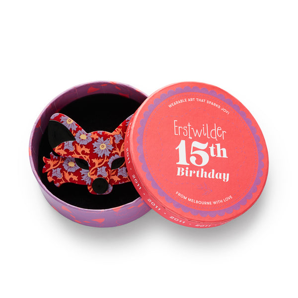 A fox face acrylic resin brooch with a red background and a floral pattern in lavender, orange, and purple, featuring solid black ear and nose areas, shown in illustrated round box packaging
