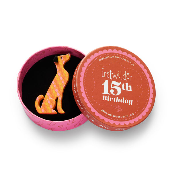 A stylized dog-shaped acrylic resin brooch featuring a geometric diamond pattern in orange, yellow, and pink, with a black nose and eyes, shown in illustrated round box packaging