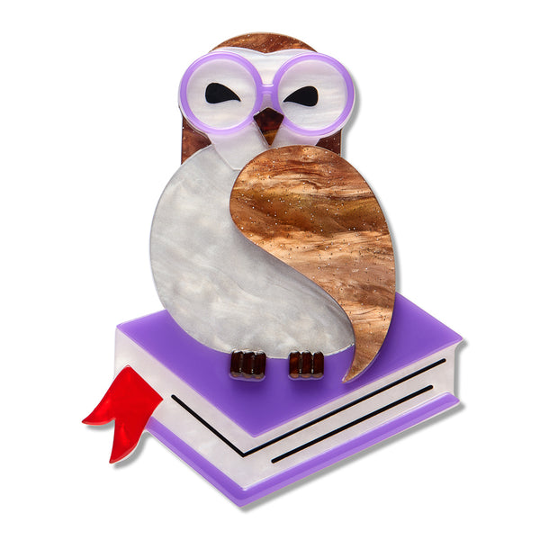 acrylic resin brooch depicting an owl wearing round purple-rimmed glasses, perched on top of a purple and white book with a red ribbon bookmark. The owl has a white body, brown head, and a marbled brown wing, set against a plain white background.