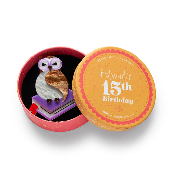 acrylic resin brooch depicting an owl wearing round purple-rimmed glasses, perched on top of a purple and white book with a red ribbon bookmark. The owl has a white body, brown head, and a marbled brown wing. Shown in illustrated round box packaging