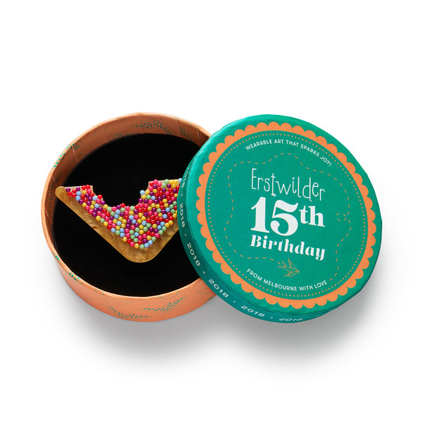 acrylic resin brooch of a triangular piece of bread covered in colorful sprinkles with a bit taken out of it, shown in illustrated round box packaging