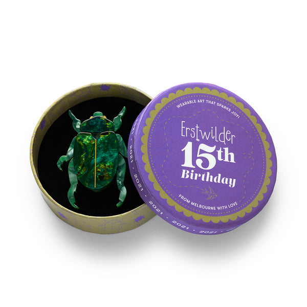 A beetle-shaped acrylic resin brooch in marbled greens with and a gold-colored central ridge, shown in illustrated round box packaging