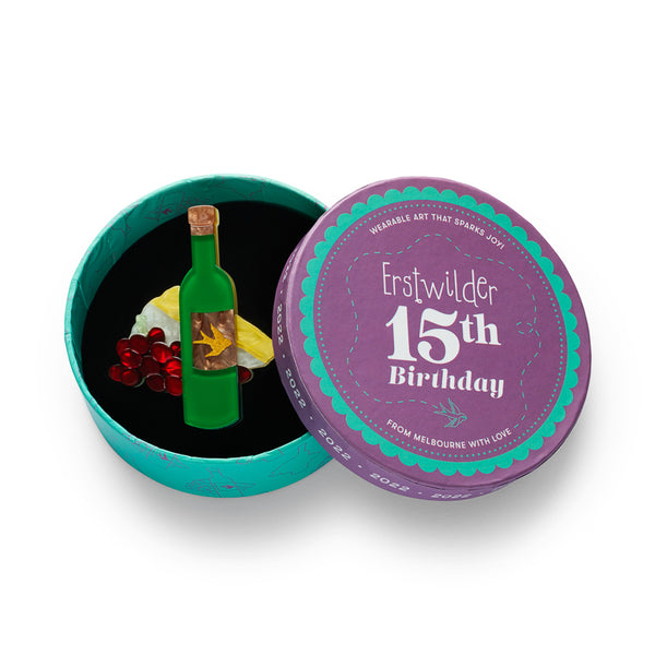 acrylic resin brooch of a green wine bottle with a cork stopper and a decorative label featuring a gold swallow on a brown background, styled with red grapes and a cheese wedge, shown in illustrated round box packaging