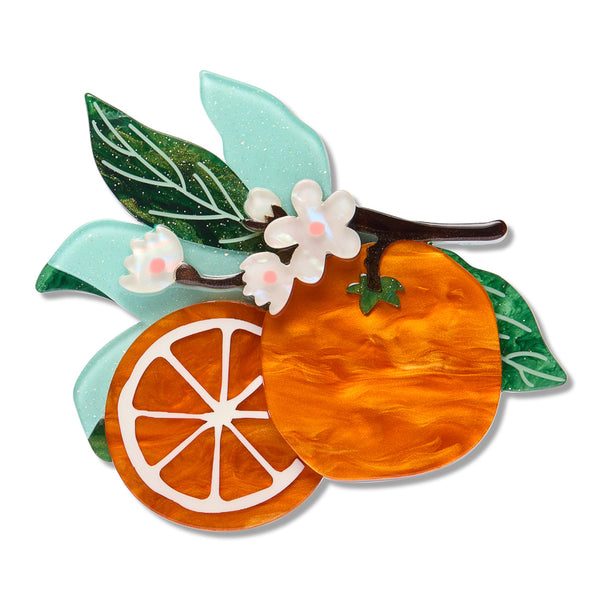 An acrylic resin fruit brooch featuring a whole orange, a sliced orange segment, green leaves, and white blossoms on a dark branch, set against a plain white background.