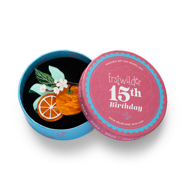 An acrylic resin fruit brooch featuring a whole orange, a sliced orange segment, green leaves, and white blossoms on a dark branch, shown in illustrated round box packaging