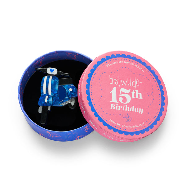 A scooter-shaped acrylic resin brooch featuring a blue marbled pattern with white stripes, a white headlight with a blue heart, and black handlebars, shown in illustrated round box packaging