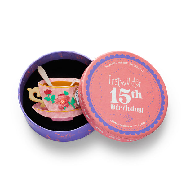 A marbled pink resin teacup and saucer set brooch with gold rim, floral decoration, a spoon inside, and a hanging tea tag reading 'Earl Grey TEA', shown in illustrated round box packaging