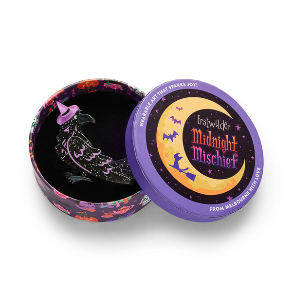 A glitter black raven acrylic resin brooch with a lavender witch hat and wing accents. Shown in illustrated round gift box packaging.