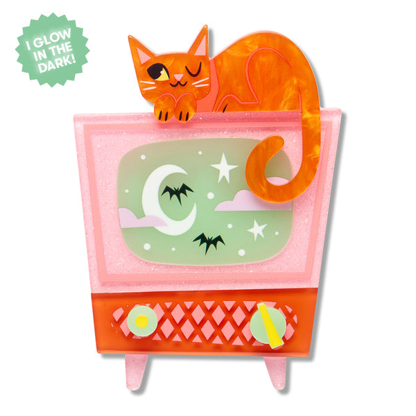 A layered acrylic resin brooch featuring an orange cat sitting on top of a pink and orange cabinet television with a glow-in-the-dark green screen showing a moon, stars, and bats, with two knobs at the bottom.