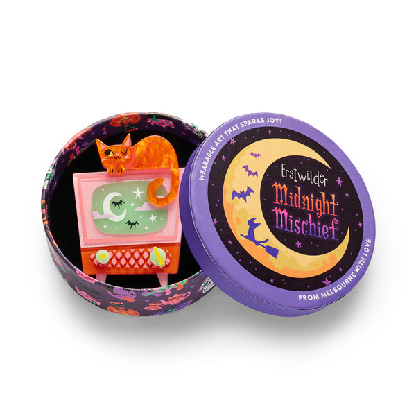 A layered acrylic resin brooch featuring an orange cat sitting on top of a pink and orange cabinet television with a glow-in-the-dark green screen showing a moon, stars, and bats, with two knobs at the bottom. Shown in illustrated round gift box packaging.