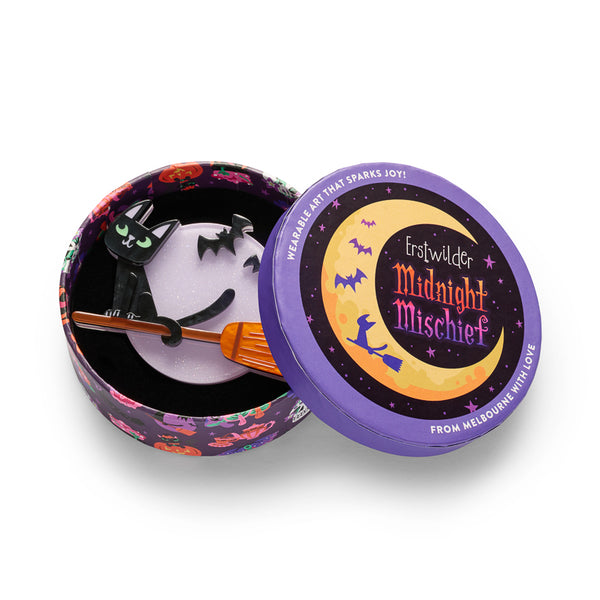 A Halloween-themed layered acrylic resin brooch featuring a black cat with green eyes sitting on an orange broomstick in front of a glittery light purple full moon, with two black bats flying above. Shown in illustrated round gift box packaging.