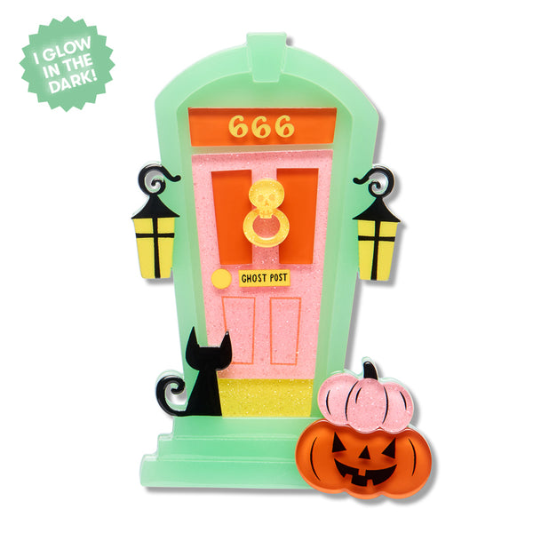 Halloween ghost house door acrylic resin brooch with a mint green frame, pink glittery door, orange number 666, black cat silhouette, yellow lanterns, and an orange jack-o'-lantern pumpkin, shown on a white background with a green 'I Glow in the Dark!' badge.