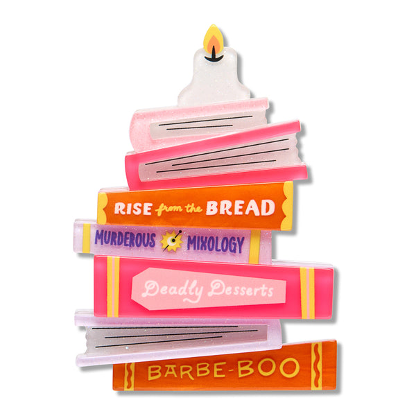 A book stack-shaped acrylic resin brooch featuring book spines in pink, orange, yellow, and white with text reading 'RISE from the BREAD', 'MURDEROUS MIXOLOGY', 'Deadly Desserts', and 'BARBE-BOO' on a plain white background.