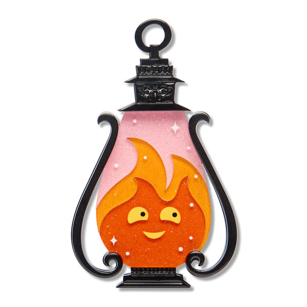 A lantern-shaped acrylic resin brooch featuring a smiling orange flame character with yellow eyes and a pink glittery interior, framed by black scroll-like sides and a black top loop, shown on a plain white background.