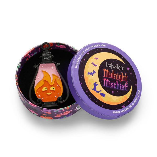 A lantern-shaped acrylic resin brooch featuring a smiling orange flame character with yellow eyes and a pink glittery interior, framed by black scroll-like sides and a black top loop, shown in illustrated round gift box packaging