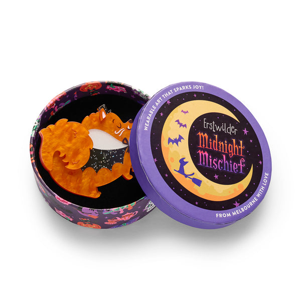acrylic resin brooch depicting the side view of a fluffy orange dog wearing a glitter black cape and top hat, and carrying a jack-o-lantern candy bucket filled with bones in its mouth. Shown in illustrated round gift box packaging.