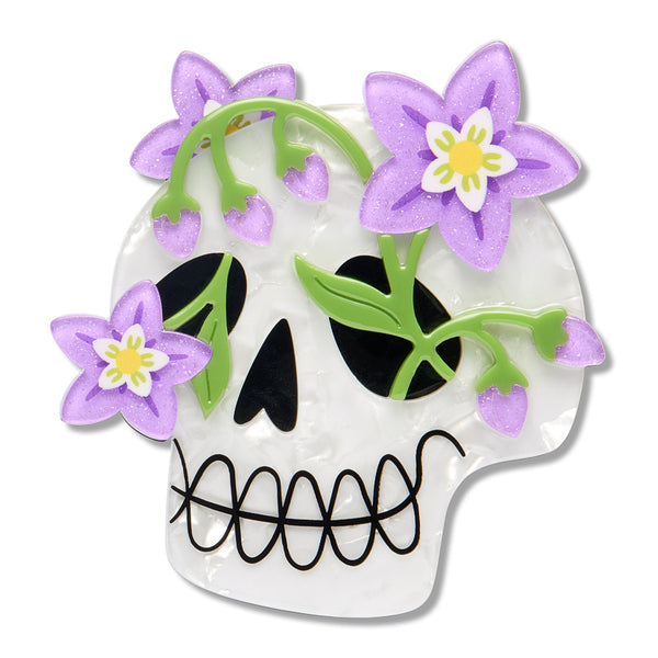 A white skull acrylic resin brooch featuring purple glitter flowers, green leaves, black eye sockets and nose, shown on a plain white background.