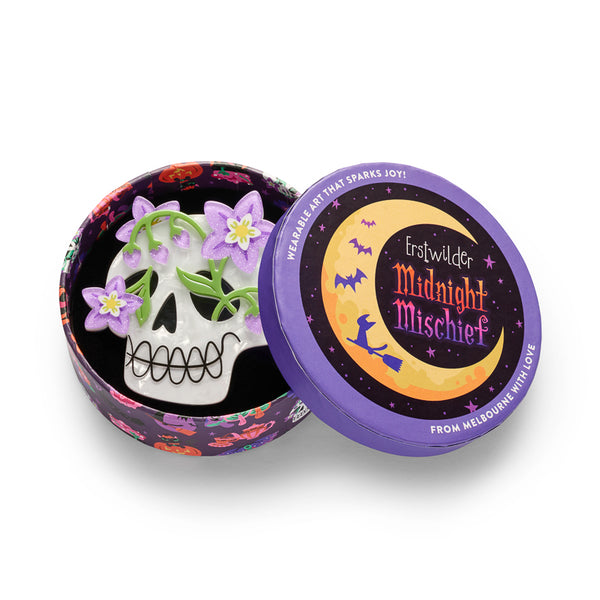 A white skull acrylic resin brooch featuring purple glitter flowers, green leaves, black eye sockets, nose, and teeth details, shown in illustrated round gift box packaging