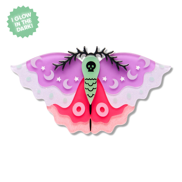 A moth-shaped glow-in-the-dark acrylic resin brooch on a white background, featuring a green thorax with a black skull motif, purple and pink wings with crescent moon and star details, and a green starburst sticker in the top-left corner that reads 'I GLOW IN THE DARK!'.