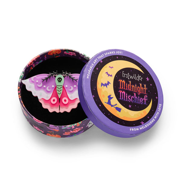 A moth-shaped glow-in-the-dark acrylic resin brooch on a white background, featuring a green thorax with a black skull motif, purple and pink wings with crescent moon and star details, shown in illustrated round gift box packaging