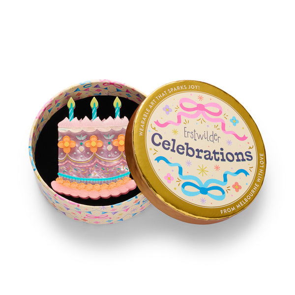 cake-shaped acrylic resin brooch with pink frosting, orange flowers, teal candles, and multicolor sprinkles. Shown in illustrated round gift box packaging.