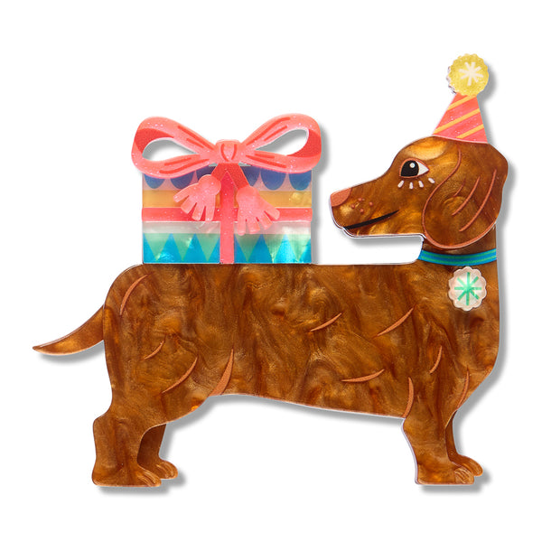 layered acrylic resin brooch shaped like a brown dachshund wearing a pink party hat, with a colorful striped gift box and pink bow on its back, shown on a white background.