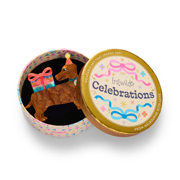 layered acrylic resin brooch shaped like a brown dachshund wearing a pink party hat, with a colorful striped gift box and pink bow on its back. Shown in illustrated round gift box packaging.