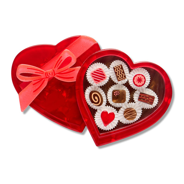 layered acrylic resin brooch of a heart-shaped red chocolate box with a matching red ribbon bow, shown open to reveal assorted decorated chocolates inside on a white background.