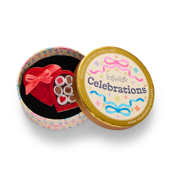 layered acrylic resin brooch of a heart-shaped red chocolate box with a matching red ribbon bow, shown open to reveal assorted decorated chocolates inside. Shown in illustrated round gift box packaging.