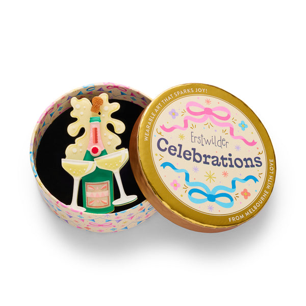 green acrylic resin champagne bottle brooch with a pink neck, two champagne glasses, and a yellow champagne splash behind it. Shown in illustrated round gift box packaging.