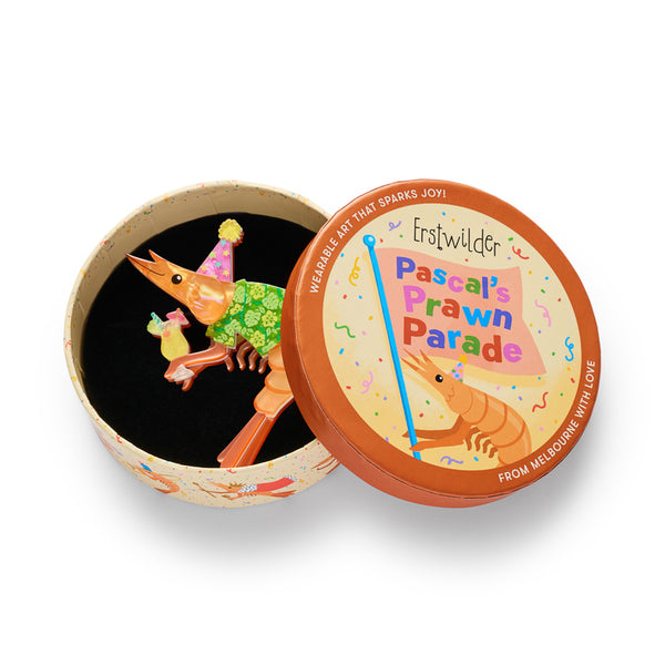 A cartoon shrimp acrylic resin brooch with an orange body, wearing a pink party hat with yellow stars and a green floral shirt, holding a tropical drink, shown in illustrated round box packaging