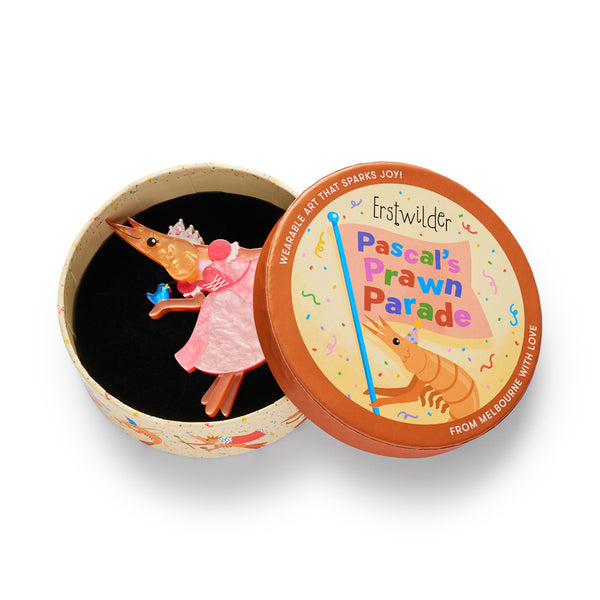 A shrimp-shaped layered acrylic resin brooch with a pink glittery dress, small tiara, and a blue bird accessory, shown in illustrated round box packaging