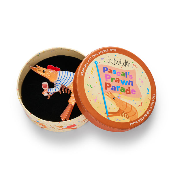 A stylized cartoon prawn layered acrylic resin brooch. The prawn is orange with a blue-and-white striped body, a red beret, and holds a pink drink in one claw. Shown in illustrated round box packaging