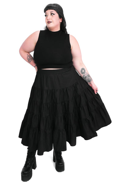Plus size model wearing a black cotton poplin midi skirt with an elastic waistband and four tiered ruffled layers with side seam pockets. Shown from front
