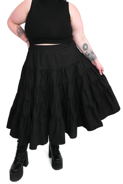 Plus size model wearing a black cotton poplin midi skirt with an elastic waistband and four tiered ruffled layers with side seam pockets. Shown from front in close up