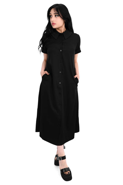 Model wearing a black rayon blend button-front slightly relaxed fit dress with a pointed collar, cuffed short sleeves, slightly a-line midi length skirt, and side seam pockets. Shown from front with hands in pockets