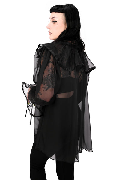 A model wearing a black chiffon sheer tunic with ruffled keyhole detail collar and 3/4 sleeves with gathered ruffled cuffs, both with black satin trim and adjustable ties. Shown from side styled with black bodysuit