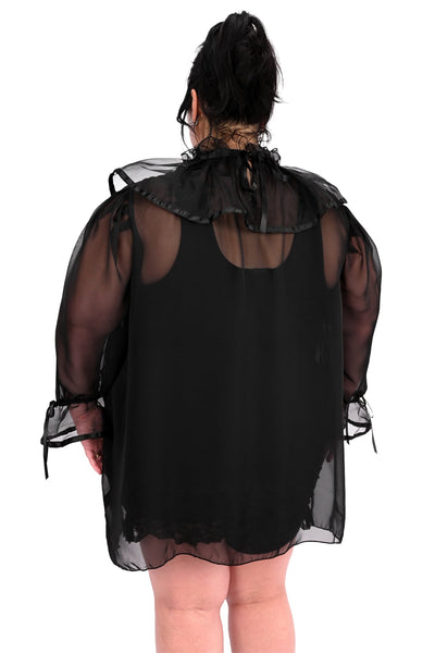 A model wearing a black chiffon sheer tunic with ruffled keyhole detail collar and 3/4 sleeves with gathered ruffled cuffs, both with black satin trim and adjustable ties. Shown from back styled with black leotard 