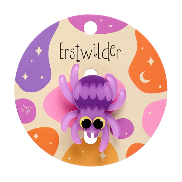 A cute cartoon spider acrylic resin brooch in shades of purple with large round black and yellow eyes, mounted on a round beige backing card decorated with purple, orange, and pink abstract shapes and the text 'Erstwilder'.