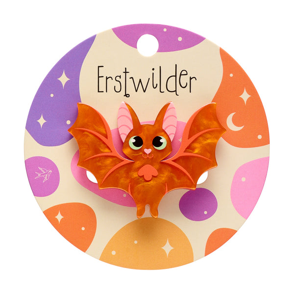 A cute cartoon bat acrylic resin brooch with orange wings and body, pink inner ears, and large green-ringed eyes, mounted on a round cream-colored backing card decorated with purple, orange, and pink celestial shapes. The brand name 'Erstwilder' is printed at the top of the card.