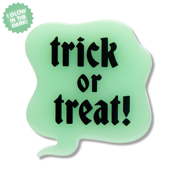 A novelty Halloween acrylic resin brooch shaped like a speech bubble with the text 'trick or treat!' in black gothic lettering on a pale mint green glow-in-the-dark material. A green starburst sticker in the top-left corner reads 'I GLOW IN THE DARK!'.