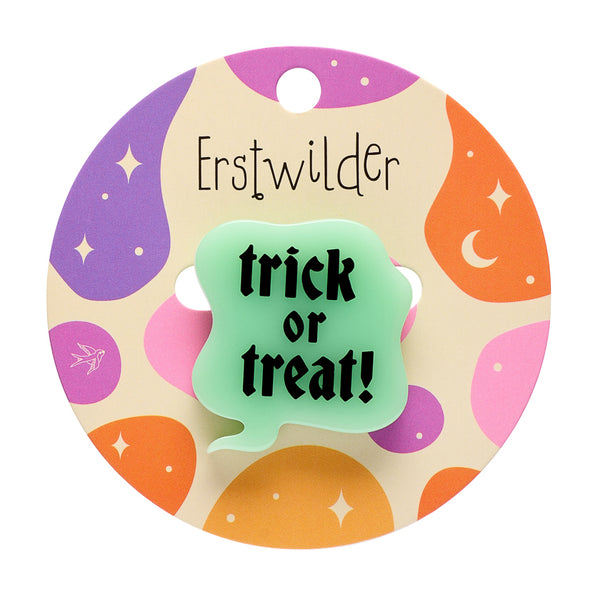 A novelty Halloween acrylic resin brooch shaped like a speech bubble with the text 'trick or treat!' in black gothic lettering on a pale mint green glow-in-the-dark material, mounted on a circular printed backer card with colorful abstract shapes in purple, orange, pink, and beige, featuring the text 'Erstwilder' 